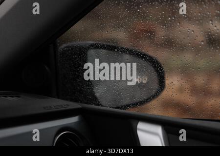 Car with lights on comes from behind on a wet country road seen through rain drops in a car side mirror, rocks & pine trees are seen too. Stock Photo