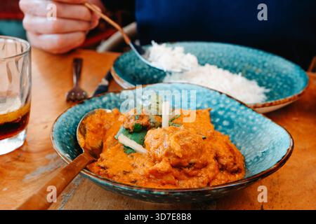a man sits at a restaurant table with a bowl of tikka masala placed in front of him, its thick orange sauce filling the dish while a serving of white Stock Photo