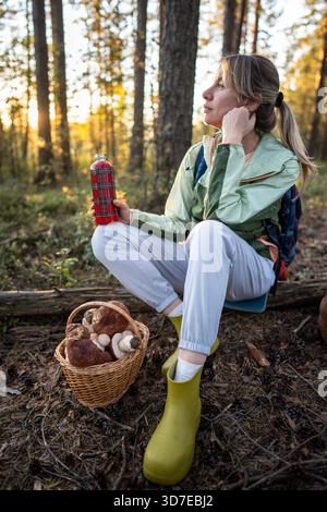 Blissful female tourist watching forest nature drink hot beverage post harvest in woodland. Quiet reflection after mushroom hunting, mindful appreciation of forest scenery, slow living on solo travel || Model released Stock Photo
