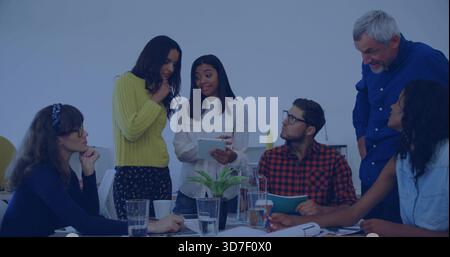 Presenting woman in yellow top holding tablet, team reviewing papers at meeting table with plant Stock Photo