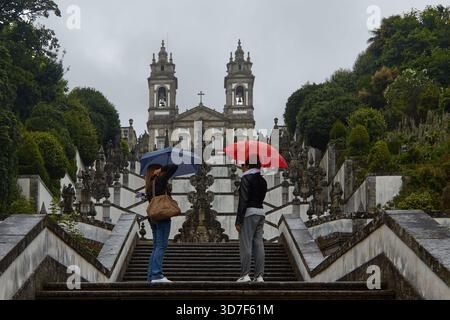 Braga, Portugal - June 12, 2025: Two girls stand on the main staircase of Bom Jesus do Monte Sanctuary, holding red and blue umbrellas on a rainy day Stock Photo