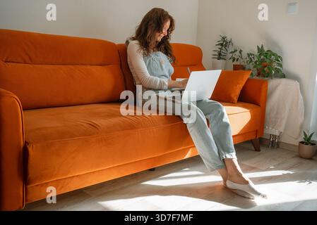 Woman with red hair sitting comfortably on an orange sofa, working or studying remotely using a laptop while taking notes, embracing a modern freelance lifestyle at home in a bright living room Stock Photo