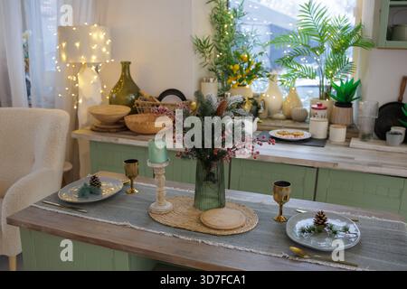 Festive Christmas kitchen with cozy dining table setting, evergreen centerpiece, candles and warm fairy lights for winter holiday home decor. Stock Photo