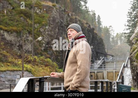 Older man in a warm sweater and scarf stands by a railing, surrounded by rocky cliffs and trees, appreciating the peaceful outdoor atmosphere and natu Stock Photo