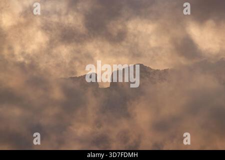 A dramatic, close-up shot of a rugged mountain peak barely visible through warm, storm-colored clouds at sunset over Benidorm. Stock Photo