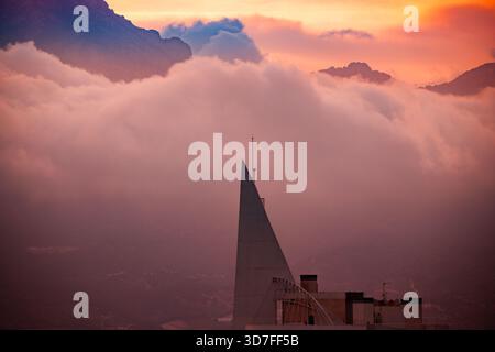 A highly contrasted and warm-toned image featuring the sharp, triangular spire of a high-rise building cutting through a massive, reddish-pink fog ban Stock Photo