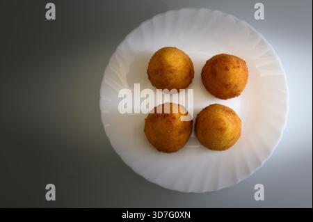 Four fried arancini are placed on a white plate. They're golden brown, ready for the fryer. The plate is set on a wooden table. Stock Photo