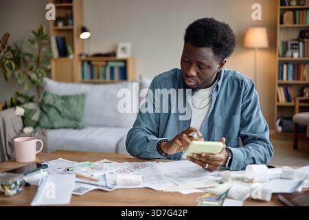 Young Black Man Reviewing Invoices and Bills Using Smartphone at Home Stock Photo
