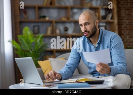 Young man analyzing financial documents and using a laptop while working from home, focusing on budgeting, bill payment, and managing remote work responsibilities Stock Photo