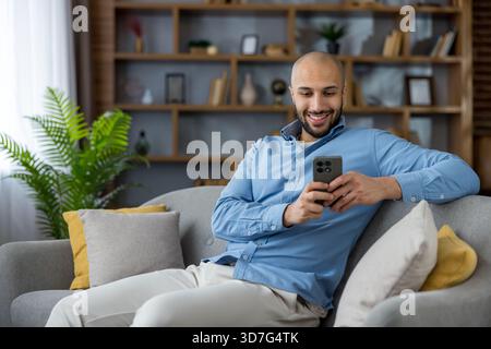 Happy young man sitting comfortably on a gray sofa at home, smiling while engaging with his mobile phone, chatting, browsing social media, and enjoying leisure time Stock Photo