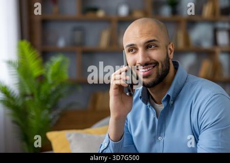Smiling bald bearded man communicating happily on a smartphone while sitting comfortably at home, enjoying a pleasant conversation and connectivity through technology Stock Photo