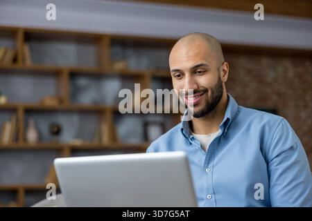 Smiling man intently looking at a laptop screen. Engaging in remote work or online communication from a casual home office setup. Representing modern digital entrepreneurship and flexible work styles Stock Photo