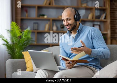 Young man with headphones on laptop in a home office, smiling during a video conference while taking notes in a notebook, focused on a remote meeting or online class Stock Photo