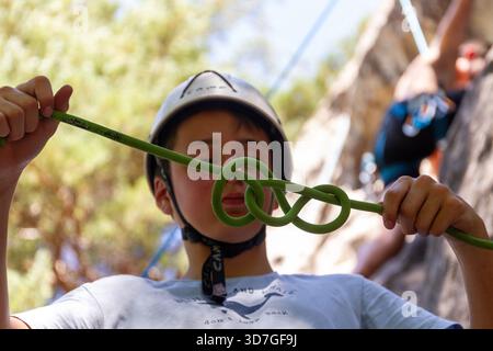 Young man rock climbing on an outdoor cliff, wearing a helmet and harness, carefully ascending the natural rock face on a sunny day. Stock Photo
