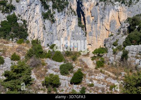 Group of hikers walking along a rocky mountain trail with steep limestone cliffs in the background on a sunny day. Stock Photo