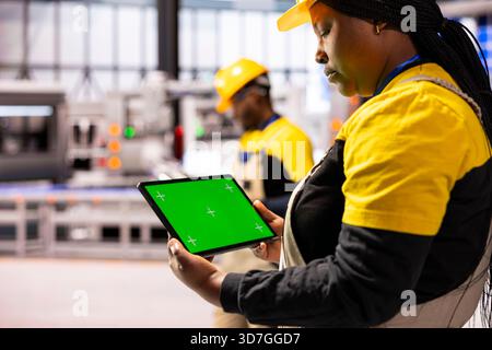Black female production engineer using tablet with blank mockup template and inspecting a factory. African American industrial manager holding green screen device, monitoring system performance. Stock Photo