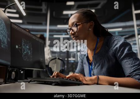 Focused data center developer using artificial intelligence deep learning tech. African american woman in server hub looking at AI programming language on PC monitor to update gear Stock Photo