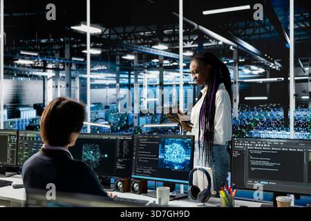 System administrators in data center troubleshoot server connectivity issues using AI. African american woman and technician in server room auditing systems for vulnerabilities Stock Photo