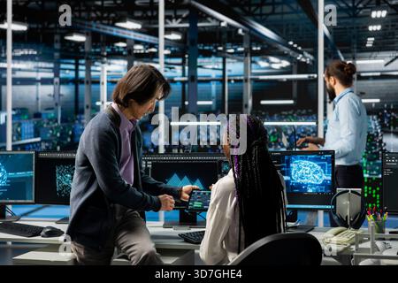 Data center manager teaching intern how to run AI forecasting simulations, tracking performance. Server hub supervisor helping trainee learn to integrate machine learning APIs into software platforms Stock Photo