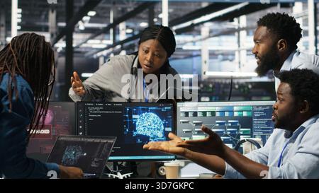 Data center engineer in wheelchair using AI requesting help from coworkers during security breach. Server farm worker with disability aided by colleagues to fight hacker attack stealing data, camera B Stock Photo