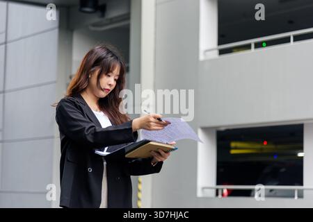 Young Asian business woman in suit busy with work, reviewing important documents and taking notes on Stock Photo