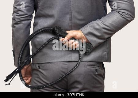 A stylish man in a gray suit holds a black whip behind his back, suggesting control, despotism, and a violently coercive form of leadership. Stock Photo
