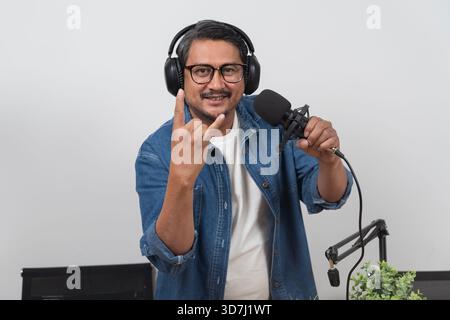 Cheerful man wearing headphones showing a rock hand sign while speaking into a microphone in a modern studio environment, representing podcasting, bro Stock Photo
