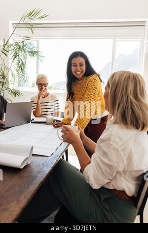 Three female architects of diverse ages collaborating on project designs in a bright workspace. One woman smiles while standing and engaging with the Stock Photo