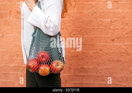 A woman holds a colorful mesh bag filled with fresh apples, standing in front of a textured orange wall. Her bright outfit contrasts beautifully with Stock Photo