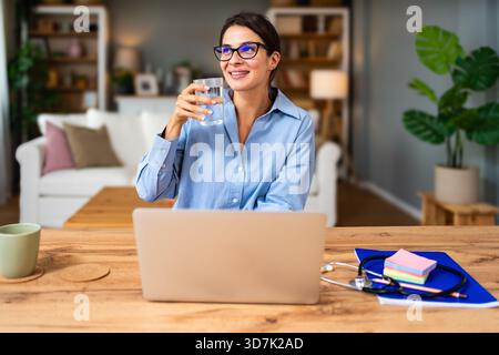 Healthcare professional conducts telemedicine session on laptop reviewing patient paperwork and typing notes while a stethoscope lies on the desk conv Stock Photo
