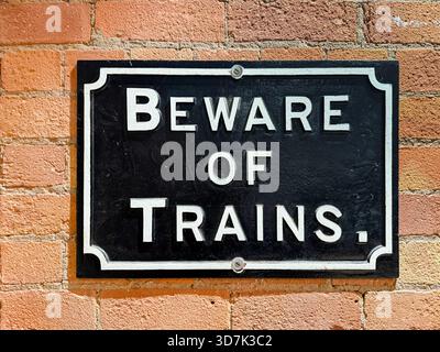 cast iron Black and white warning sign reading 'Beware of Trains' mounted on a red brick wall near a railway or station area Stock Photo