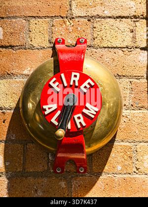Close-up of a vintage red and brass fire alarm bell mounted on a brick wall under direct sunlight Stock Photo