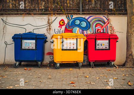Three trash cans are lined up against a wall with graffiti on it. The cans are blue, yellow, and red Stock Photo