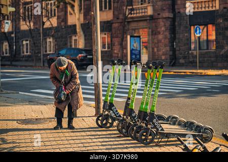 Yerevan Armenia 11.20.2025. A senior old woman is standing next to a row of scooters. The scooters are green and are parked on the sidewalk Stock Photo