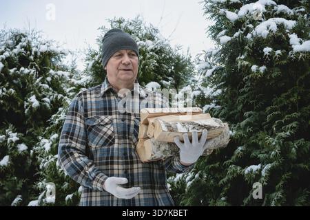 Senior man carrying firewood outside in snowy backyard. Practical winter preparation for home heating. Realistic lifestyle image for energy, fuel, and Stock Photo