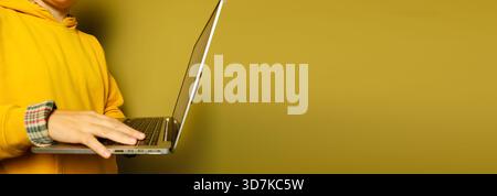 Person in yellow hoodie typing on silver laptop against mustard yellow background, cropped modern style. Online banner for design with copy space Stock Photo