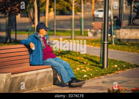 Yerevan Armenia 11.20.2025. An older man sits on a bench in a park. He is wearing a blue jacket and a hat. The park is empty, and there are no other p Stock Photo