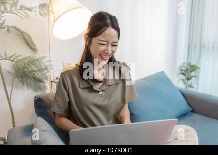 A young woman sits on a sofa with a laptop, smiling while looking at the screen. She appears relaxed and engaged, enjoying online communication, remot Stock Photo