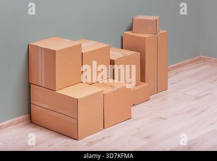 Moving boxes are stacked in the corner of a room with a light wood floor and a grey wall Stock Photo