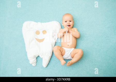 Cute baby lies with a toothbrush on a blue background. White tooth made of fabric. Medicine, dentistry, health concept || Model released Stock Photo
