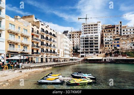 Xlendi, Gozo, Malta - April 12, 2025: Vibrant waterfront scene in Xlendi Bay with outdoor cafes, colorful jet skis and modern buildings rising behind traditional limestone houses Stock Photo