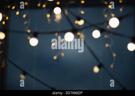 Soft defocused string lights glowing against dark blue evening background. Warm bokeh bulbs, festive mood. Abstract blurred illumination for seasonal, Stock Photo
