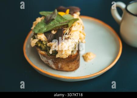 Close-up of a gourmet sourdough toast topped with creamy scrambled eggs, fresh avocado, and greens, served on a stylish plate against a dark table background, ideal for breakfast and trendy cafe concepts Stock Photo