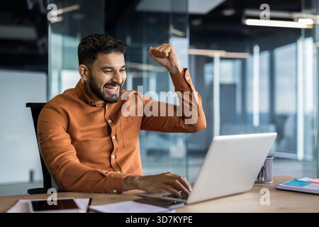 Successful indian businessman raising a fist in victory, celebrating professional achievement or good news while working on a laptop at an office desk Stock Photo