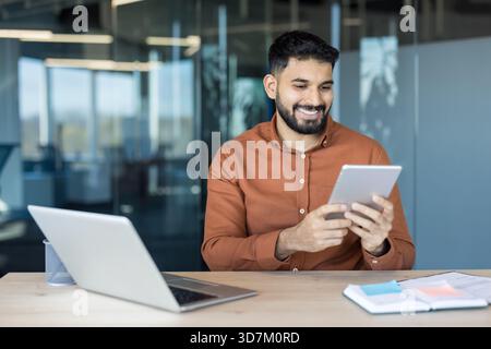 Smiling young professional man with a beard sitting at his desk, cheerfully using a digital tablet while a laptop and planner are on the table, indicating a modern and efficient work environment Stock Photo