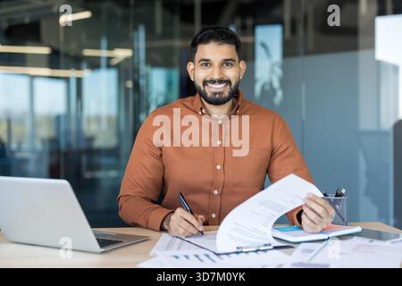 Indian businessman smiling at camera, sitting at his office desk, working with a laptop, signing financial documents, and reviewing charts and reports Stock Photo