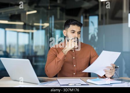 Focused businessman sitting at office desk, thoughtfully analyzing documents and reviewing data while working on a laptop, concentrating on a new business strategy Stock Photo