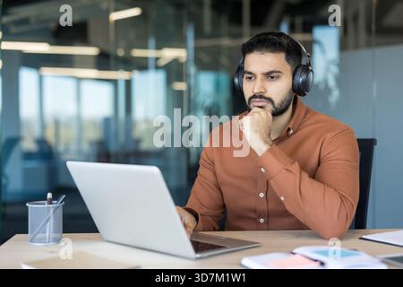 Young man wearing headphones thoughtfully working on a laptop in a modern office, engaged in focused online learning or virtual meeting while observing the screen Stock Photo