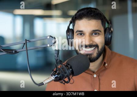 Young indian man in a brown shirt smiling while wearing headphones and speaking into a professional microphone, actively creating audio content or broadcasting in a studio setting Stock Photo