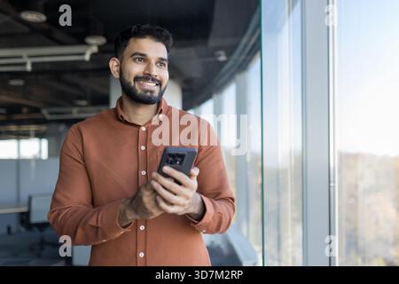 Young indian businessman smiling, enjoying connection and success, standing in a contemporary office while holding a smartphone and looking out a large window Stock Photo